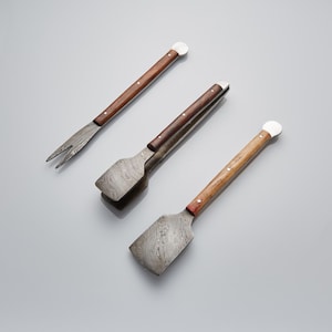 Puede incluir: Juego de tres utensilios para barbacoa. El juego incluye un tenedor, unas pinzas y una espátula. Cada herramienta presenta una cabeza de metal estampada y un mango de madera con una tapa blanca en el extremo. Los mangos son de color marrón medio.
