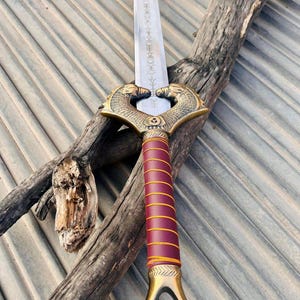 K&ouml;nnte beinhalten: Ein silbernes Schwert mit einem goldfarbenen Griff und einem rot-goldenen Griff. Das Schwert hat ein Doppelkopf-Schlangen-Design am Griff und ein dekoratives Muster auf der Klinge.