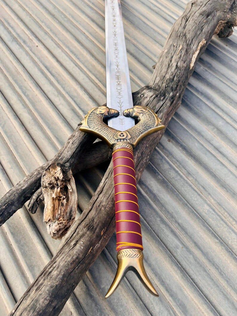 K&ouml;nnte beinhalten: Ein silbernes Schwert mit einem goldfarbenen Griff und einem rot-goldenen Griff. Das Schwert hat ein Doppelkopf-Schlangen-Design am Griff und ein dekoratives Muster auf der Klinge.