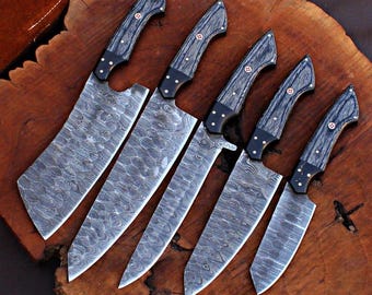 Puede incluir: Conjunto de cinco cuchillos de cocina con mangos de madera negra y hojas de acero Damasco. Las hojas tienen un patrón ondulado y están ordenadas por tamaño.