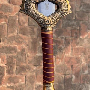 K&ouml;nnte beinhalten: Ein Schwert mit einem goldfarbenen Griff, der mit zwei stilisierten Tierk&ouml;pfen verziert ist. Der Griff ist mit burgunderfarbenem Leder mit goldenen Akzenten umwickelt. Die Klinge ist silberfarben und hat ein dekoratives Design.