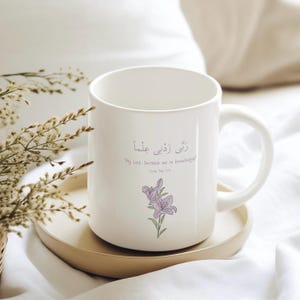 May include: White ceramic mug with a purple flower design and the Arabic text "رزقني علماً" which translates to "My Lord, increase me in knowledge." The text is in smaller font below the flower design and reads "Surah Taha 114".