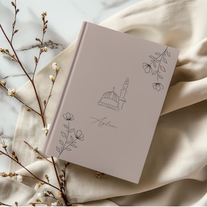 Pode incluir: Um caderno marrom claro com uma ilustração de uma mesquita e a palavra "Asylan" escrita em itálico. O caderno é decorado com desenhos florais simples.