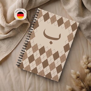 May include: A spiral-bound notebook with a tan argyle pattern and a brown Arabic letter on the cover. The notebook is open, and the pages are visible. The background is a neutral beige fabric.