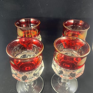 May include: Set of four red glass wine glasses with a clear glass stem and base. The glasses have a textured design and a gold rim.