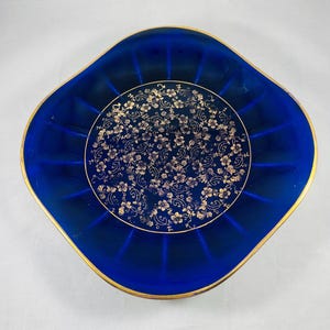 May include: A vintage, square-shaped, cobalt blue dish with a gold rim. The center features a gold floral pattern. The dish has a ribbed design radiating from the center. A decorative piece for serving or display.