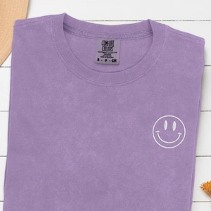 Könnte beinhalten: Ein lavendelfarbenes T-Shirt mit einem weißen Smiley, der auf die linke Brust gestickt ist. Das Shirt ist aus weichem, bequemem Stoff und hat eine lockere Passform.