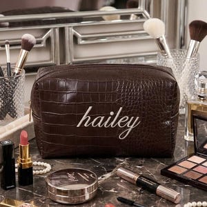 May include: A dark brown rectangular cosmetic bag with a crocodile-embossed texture and the name "hailey" embroidered in white script on the front. The bag features a top zipper closure and is constructed from a structured, durable material.