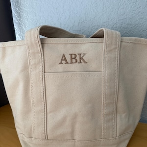 Könnte beinhalten: Eine beigefarbene Canvas-Tragetasche mit genähten Nähten und Griffen. Die Initialen "ABK" sind in einem dunkleren beigefarbenen Faden auf der Vordertasche gestickt. Die Tasche ist zum Tragen von Gegenständen konzipiert.
