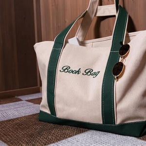 May include: A cream-colored canvas tote bag with dark green trim and handles. The words "Book Bag" are embroidered in dark green script on the front. A pair of round, gold-rimmed sunglasses with dark lenses is hooked onto one of the green straps.