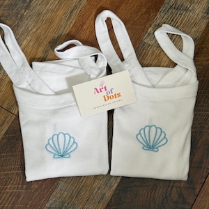 Puede incluir: Dos bolsas de mano blancas con un diseño de concha de color azul claro bordado en la parte delantera. Las bolsas tienen correas blancas y se exhiben sobre una superficie de madera. Una etiqueta con el texto "Art of Dots EMBROIDERED APPAREL" es visible.