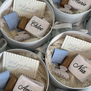 May include: Round gift boxes with striped sides containing a card reading Will you be my bridesmaid or maid of honor, a light blue pouch, a brown tube, a white headband embroidered with a name, and a patterned hair claw clip.