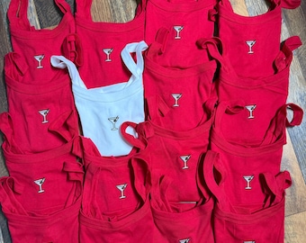 May include: A collection of red tank tops with a small embroidered martini glass design. One white tank top is also present. The tank tops have a scoop neck and tie straps. The tops are likely for a party or event.