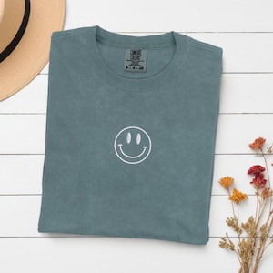 Op de afbeelding: Een tealblauw t-shirt met een wit geborduurd smiley-gezicht. Het shirt is gevouwen en ligt plat op een wit houten oppervlak.