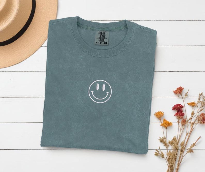 Op de afbeelding: Een tealblauw t-shirt met een wit geborduurd smiley-gezicht. Het shirt is gevouwen en ligt plat op een wit houten oppervlak.