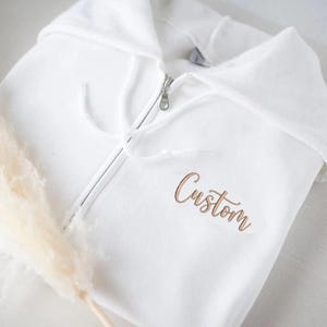Puede incluir: Una sudadera blanca con cremallera y capucha con cordón. La palabra "Custom" está bordada en cursiva en la parte delantera. La cremallera es plateada. Una ramita de hierba de la pampa seca está en primer plano.