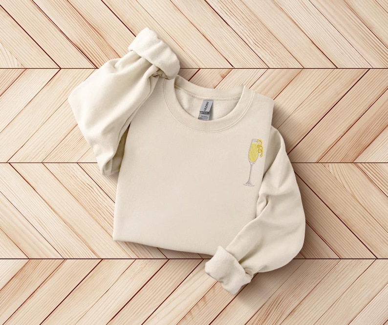 K&ouml;nnte beinhalten: Cremefarbenes Sweatshirt mit einem kleinen, gestickten Mimosa-Glas-Design auf der Vorderseite. Das Mimosa-Glas ist gelb und wei&szlig;. Die &Auml;rmel sind hochgekrempelt. Das Sweatshirt ist aus weichem Material.