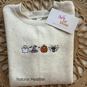 Könnte beinhalten: Ein Sweatshirt in Natur-Heather mit gestickten Halloween-Motiven: ein Geist, ein Hexenhut, ein Kürbis und eine Fledermaus. Der Text "Natural Heather" ist unten aufgedruckt. Ein Etikett mit dem Text "Art of Dots EMBROIDERED APPAREL" ist angebracht.