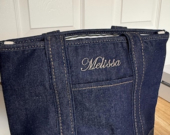 May include: A dark blue denim tote bag featuring thick, tan-colored contrast stitching along the seams and handles. The front of the bag displays the name Melissa embroidered in a white cursive script font, centered above a small open pocket.