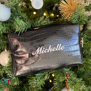 May include: A black rectangular cosmetic bag with a glossy crocodile skin texture. The name Michelle is embroidered in white cursive script across the center of the front panel. The bag features a gold-toned zipper pull on the right side.