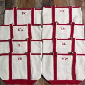 May include: A collection of white canvas tote bags with red trim and handles. Each bag has a single pocket and a monogram in the center. The initials on the bags are: KC, KW, HF, EB, EO, LM, BL, and MW.