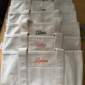 May include: A stack of beige canvas tote bags, each with a different name embroidered in a contrasting color. Names include Alayna, Sam, Eva, Alexis, Ashley, and Lauren. The bags have a front pocket and sturdy handles.