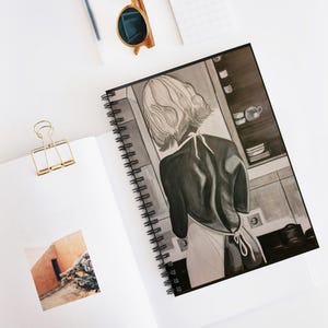 Puede incluir: Un cuaderno de espiral con una ilustración en blanco y negro de una persona de pelo corto con delantal en una cocina. La portada muestra una escena de cocina con estantes, vajilla y enchufes. Se incluye un clip de encuadernación dorado.