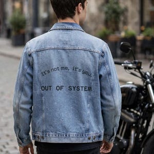 Puede incluir: Una chaqueta de mezclilla azul claro con cuello clásico, mangas largas y puños abotonados. La parte posterior presenta un texto impreso en negro que dice "It's not me, it's you." seguido de "OUT OF SYSTEM" en una fuente sans-serif.