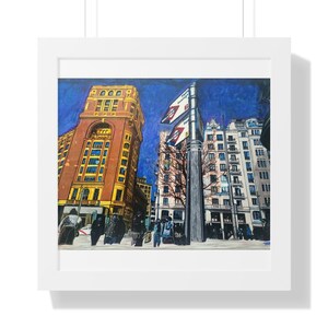Puede incluir: Una impresión artística de una escena de calle urbana, enmarcada en blanco. La obra representa edificios en tonos marrones, amarillos y blancos, con un cielo azul profundo. Se ven peatones en la calle y banderas en un poste.