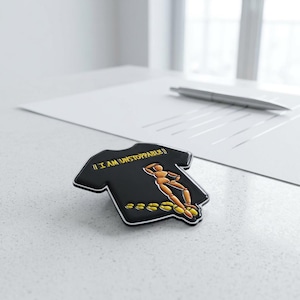 Puede incluir: Un pin de esmalte negro con forma de camiseta que presenta una figura de maniquí de madera y huellas amarillas. El texto "I AM UNSTOPPABLE" está impreso en amarillo sobre la figura. El pin tiene un borde blanco y un acabado brillante.