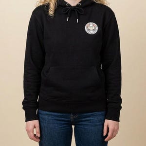 May include: A black long-sleeve hooded sweatshirt featuring a drawstring hood with metal aglets and a circular graphic on the upper left chest. The graphic depicts a crab illustration with the text "NO SIERRA" printed in a circular border.