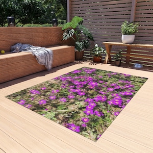 Puede incluir: Una alfombra rectangular que presenta una impresión fotográfica de alta resolución de flores púrpuras vibrantes con centros amarillos dispersas sobre un campo de hierba verde, exhibida sobre una terraza de madera clara.