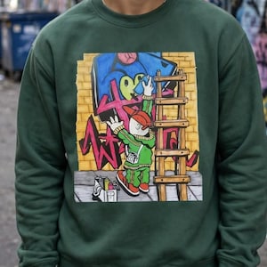 Puede incluir: Una sudadera de cuello redondo de color verde oscuro con un estampado gráfico de un personaje de dibujos animados con gorra roja y chándal verde, subido a una escalera mientras pinta con spray un muro de ladrillo amarillo.