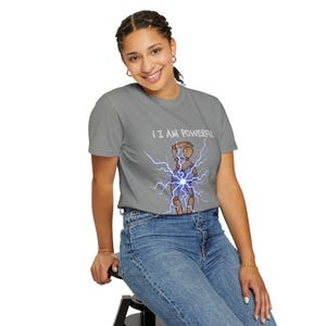 Puede incluir: Una camiseta gris con el texto "I AM POWERFUL" sobre un gráfico de una figura de madera con rayos azules. La persona de la imagen lleva vaqueros azules.