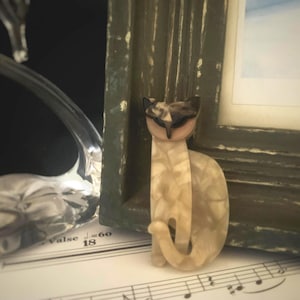 Op de afbeelding: Decoratieve kattenfiguur met een crème- en bruin gemarmerd patroon. De kat staat naast een verweerde groene fotolijst. Een heldere glazen zwaan en bladmuziek zijn op de achtergrond.