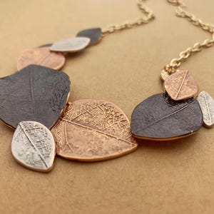 May include: A gold-coloured necklace with leaf-shaped pendants in bronze, silver, and dark brown. The detailed leaves create a nature-inspired jewellery piece. The chain is gold-coloured.