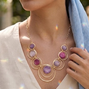 Könnte beinhalten: Eine goldfarbene Halskette mit einer Reihe von miteinander verbundenen kreisförmigen Metallringen und glatten, abgerundeten Cabochon-Steinen in verschiedenen Violett-, Magenta- und Hellrosatönen, die an einer feinen goldfarbenen Gliederkette hängt.