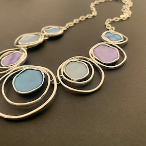 May include: Silver necklace with a chain and various pendant designs. The pendants have a circular frame with a coloured, oval centre. Colours include shades of blue and purple.