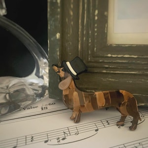 Op de afbeelding: Een teckelbeeldje met een zwarte hoge hoed. De hond is bruin en bruin met een gestreept patroon. Het beeldje staat op een bladmuziek met een ingelijste foto op de achtergrond.