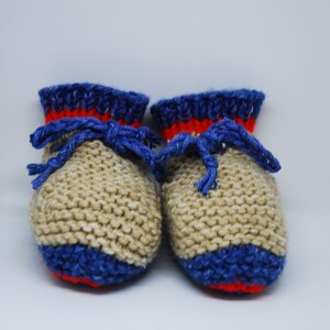 May include: A pair of knitted baby booties with a beige body, blue and red trim, and blue laces.