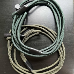 Pode incluir: Dois comprimentos de corda tran&ccedil;ada enrolados, um em verde floresta profundo e o outro em verde oliva suave. Cada corda apresenta pontas cil&iacute;ndricas pretas lisas e um mecanismo deslizante de pl&aacute;stico preto para ajustar o comprimento do cord&atilde;o.