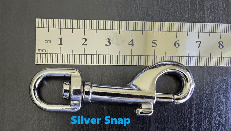May include: A silver-coloured metal swivel snap hook positioned below a metal ruler. The snap hook measures approximately 8.2 cm in total length. The text "Silver Snap" is printed in blue below the metal hardware component.