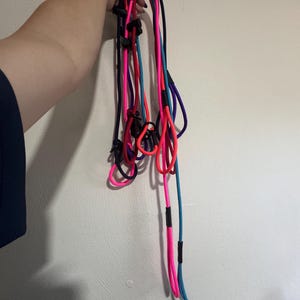 K&ouml;nnte beinhalten: Eine Ansammlung mehrerer langer, d&uuml;nner und flexibler Schn&uuml;re in leuchtendem Neonpink, hellem Blau, Dunkelviolett und Schwarz. Jede Schnur verf&uuml;gt &uuml;ber verstellbare Schieber und Verbindungsst&uuml;cke aus schwarzem Kunststoff, die Schlaufen bilden.