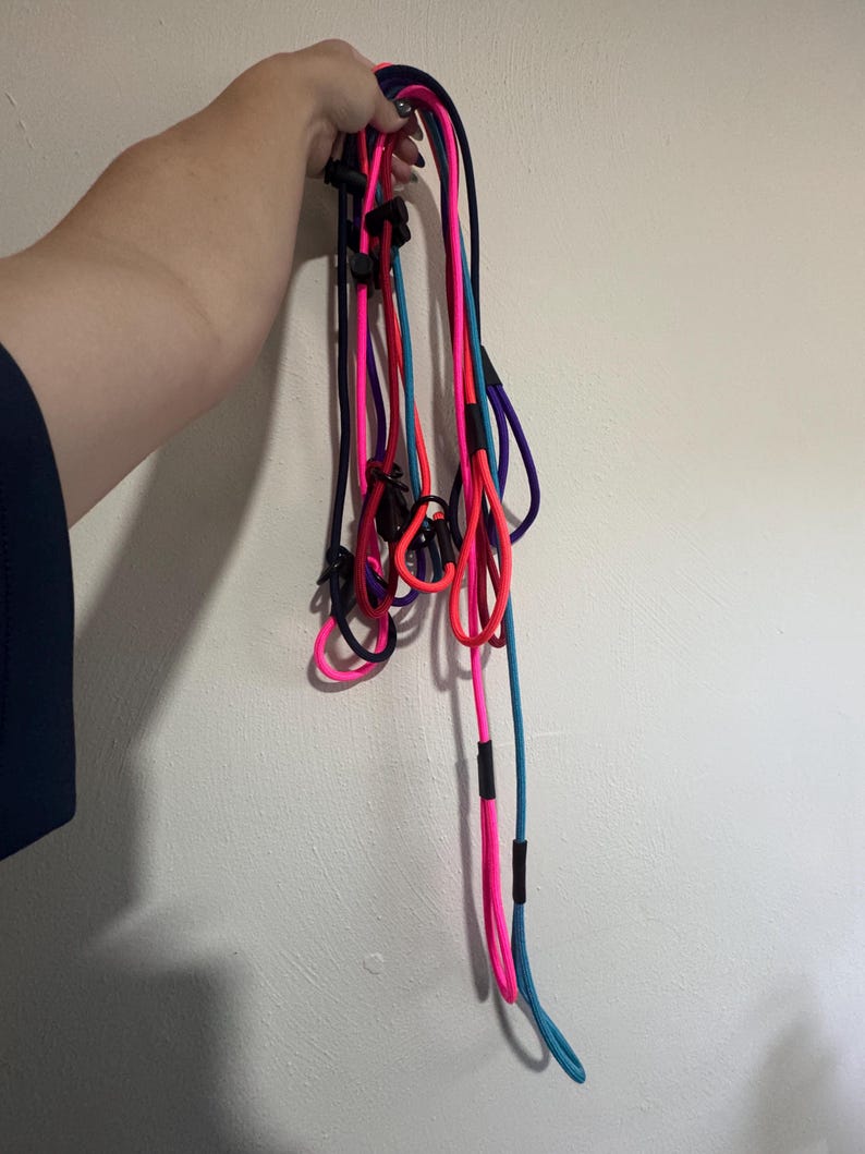 K&ouml;nnte beinhalten: Eine Ansammlung mehrerer langer, d&uuml;nner und flexibler Schn&uuml;re in leuchtendem Neonpink, hellem Blau, Dunkelviolett und Schwarz. Jede Schnur verf&uuml;gt &uuml;ber verstellbare Schieber und Verbindungsst&uuml;cke aus schwarzem Kunststoff, die Schlaufen bilden.