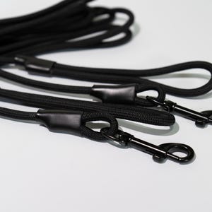 May include: A set of black rope leashes featuring durable braided cord construction with matching black metal swivel snap hooks. Each leash includes a reinforced black rubberized connector sleeve at the handle and the attachment point.