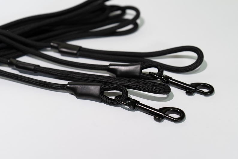 May include: A set of black rope leashes featuring durable braided cord construction with matching black metal swivel snap hooks. Each leash includes a reinforced black rubberized connector sleeve at the handle and the attachment point.
