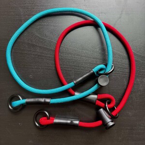 May include: Two braided rope leads, one turquoise and one red, each featuring black metal rings at the ends and black plastic sliding adjusters. The ropes are constructed with a woven texture and secured with black heat-shrink tubing at the connection points.