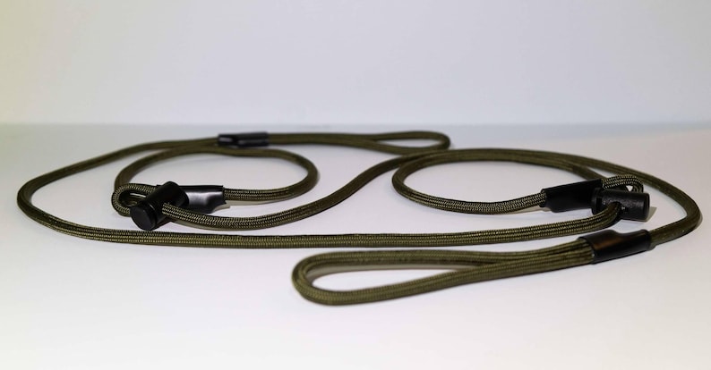 Pode incluir: Uma trela de corda verde-azeitona com dois la&ccedil;os ajust&aacute;veis e deslizadores de pl&aacute;stico preto. O cord&atilde;o &eacute; feito de um material entran&ccedil;ado e o design inclui v&aacute;rios la&ccedil;os integrados para um manuseamento seguro e pontos de fixa&ccedil;&atilde;o.