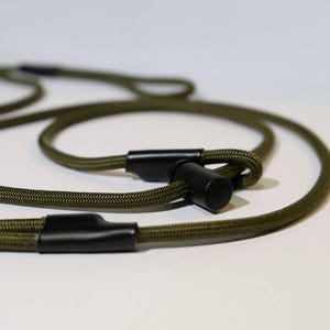 May include: Olive green dog leash with black connectors. The leash is made of a durable, woven material and features a loop handle. The black connectors are made of a smooth, solid material. The leash is designed for pet owners.