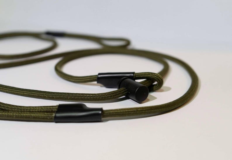 Pode incluir: Uma trela de corda verde-oliva com uma textura tran&ccedil;ada suave. A trela inclui componentes de hardware cil&iacute;ndricos pretos que prendem as al&ccedil;as da corda, proporcionando um design funcional com uma constru&ccedil;&atilde;o de cord&atilde;o tecido dur&aacute;vel.